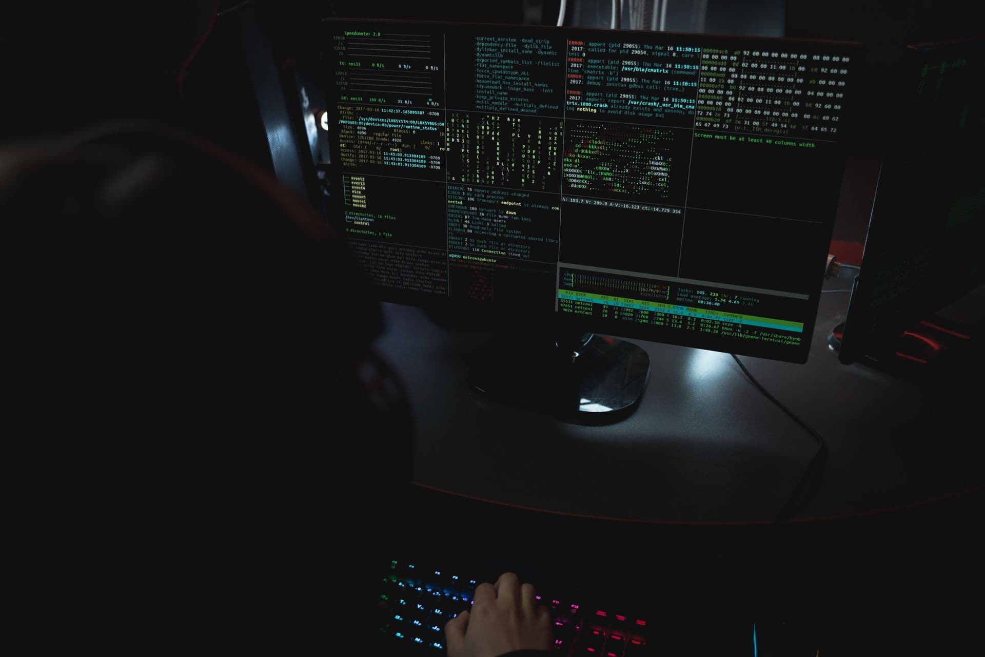 A dark room lit by monitors running code — evoking the attacker dashboard behind Kali365, which tracks targets in real time while they sign in to the real Microsoft login page.